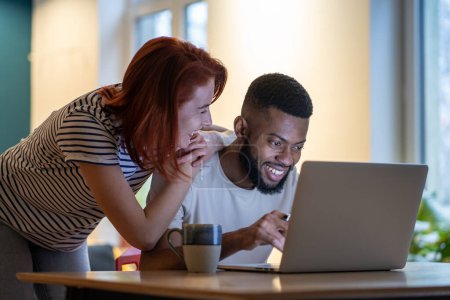 Photo for Loving young caucasian woman talking with boyfriend working on laptop late at home, smiling girlfriend demanding attention from busy with work African American guy freelancer. Work life balance - Royalty Free Image