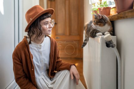 Photo for Interested woman cat owner in hat looks to fluffy pet lying on radiator under sunny window. Interaction domestic animal and mistress. Good attitude to environment joy of home. Inspiration from caring. - Royalty Free Image