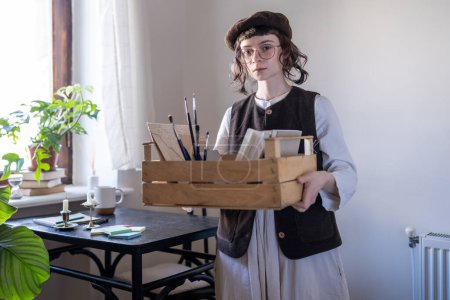 Photo for Designer illustrator girl preparing for freelance drawing order looking at camera. Creative artist young woman in stylish beret holds wooden box with art tools, materials, paints for drawing at home. - Royalty Free Image