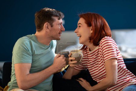 Téléchargez les photos : Joyeux couple marié rire ensemble matin avec tasse de café assis sur le canapé. Femme souriante avec mari profiter des vacances à la maison. Joyeux week-end en famille européenne amusez-vous raconter les nouvelles, blagues. - en image libre de droit