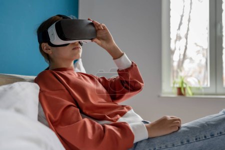 Foto de Niña adolescente relajada en gafas vr viendo video educativo inmersivo de 360 grados. Mujer con gafas VR usando auriculares metaversos acostada en la cama en casa. Realidad virtual, niños y educación - Imagen libre de derechos