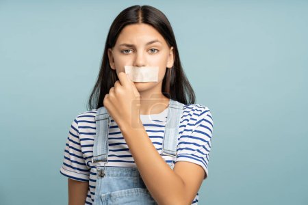 Photo for Sad frustrated teen girl rips off tape from mouth on turquoise background looking at camera. Silence, concealment of facts, domestic violence, diet, eating disorders, weak women, teenagers rights. - Royalty Free Image