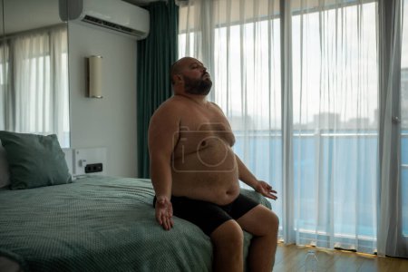 Photo for Overweight man in pants sitting in bed in bedroom with closed eyes, suffering from hot weather, trying to restore breathing, meditating. Obesity male, health problems, plump chubby tired young guy. - Royalty Free Image