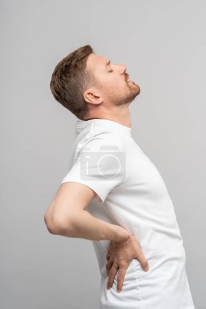 Photo for Man suffers from spinal osteochondrosis touches back on grey background. Painful facial expression with closed eyes. Sedentary work, scoliosis, lower back pain, hernia, sedentary lifestyle concept. - Royalty Free Image