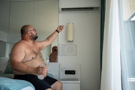 Téléchargez les photos : Homme en surpoids allume le climatiseur dans la chambre d'hôtel pour se rafraîchir par temps chaud en vacances. Gros gars avec torse nu souffre de chaleur dans un climat tropical humide. Coup de chaleur, concept de surchauffe. - en image libre de droit