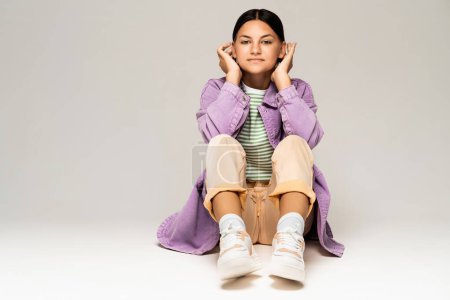 Photo for Teen girl smiling sitting on floor in stylish clothes on light studio gray background. Tanned trendy relaxed teenager looking at camera with copy space for advertising. - Royalty Free Image