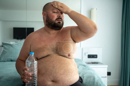 Photo for Fat man with water bottle in hand suffering from heat in hotel room on vacation in summer hot weather. Overweight bearded balding middle aged male has headache, high blood pressure, health problems. - Royalty Free Image