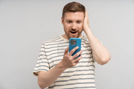 Photo for Shocked man looking at smartphone screen clutching his head on grey background. Confused amazed bearded guy losing money, having problems bad news, feeling frustration. Stress situation, life trouble. - Royalty Free Image