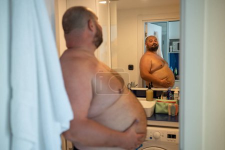 Photo for Overweight smiling man with naked torso suffering from extra weight looking at mirror touching abdomen in bathroom at home. Bearded middle aged male inspecting body to change unhealthy life habits. - Royalty Free Image