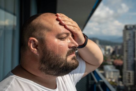 Photo for Portrait overweight man has headache touching forehead in heat summer weather. Bearded guy with excess weight looking from balcony on street in city. Extra high temperature, health problems concept. - Royalty Free Image