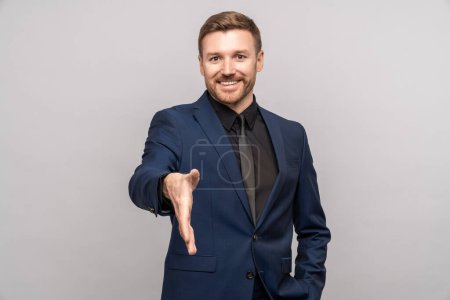 Photo for Happy businessman holds out hand for handshake looking at camera on gray background. Man in suit smile greeting somebody hand gesture. Positive friendly successful guy on meeting. Business etiquette. - Royalty Free Image