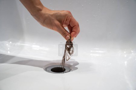 Photo for Water drain hole in bath is clogged with hair clump in bathroom at home. Woman cleaning sewer trap taking hairs, hand close-up. Bath plug hole blocked with dirt. Problem with shower sewerage. - Royalty Free Image