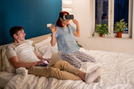 Photo for Woman in VR headset on bed, man watching, laughing, smiling. Married romantic couple, happy fun joyous cheerful pastime in evening playing in virtual reality. Satisfied girl uses VR for entertainment - Royalty Free Image