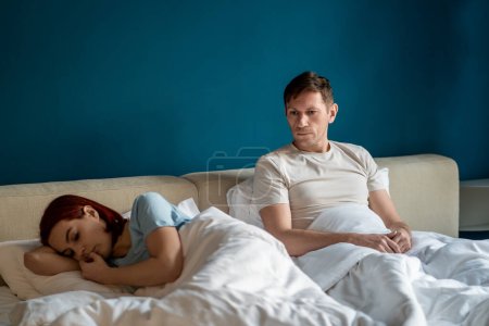 Photo for Unhappy couple ignores each other after major quarrel. Offended girl lying on side, not talking to guy sitting on bed. Misunderstandings in relationships, conflicts, unspoken grievances, sadness. - Royalty Free Image