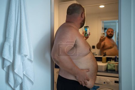 Photo for Overweight man with naked torso taking selfie on smartphone looking at mirror in bathroom at home. Middle aged male photographing himself on mobile phone for social media. Obesity, fat, chubby guy. - Royalty Free Image