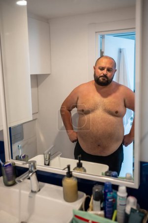 Photo for Sad overweight man dissatisfied with body shape looking at mirror in bathroom at home. Obesity, overweight male, extra weight, healthy problems, weight control. Fat guy suffering from bad habits. - Royalty Free Image