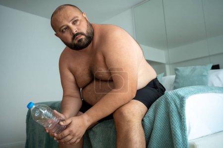 Téléchargez les photos : Gros homme assis sur le lit avec une bouteille d'eau. Jeune barbu mécontent obèse gars ne peut rassembler la force de faire des tâches quotidiennes en raison de la puissance de succion et la motivation chaude chaleur d'été provoque la paresse. - en image libre de droit