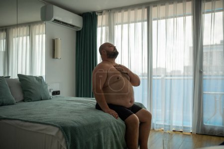 Photo for Fat middle aged man does exercises for breath to cope with summer stuffiness. Overweight obese chubby young male sitting on bed, difficult breathing deeply to handle with terrible heat, holding chest - Royalty Free Image