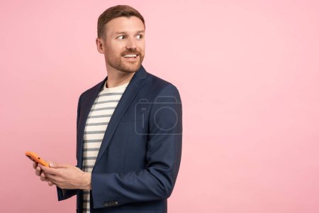 Téléchargez les photos : Homme souriant en costume veste avec smartphone dans les mains regardant l'espace de copie sur fond rose. Bannière publicitaire, affiche. Un type positif tient son téléphone portable. Homme d'affaires, pigiste, commerçant, investisseur. - en image libre de droit