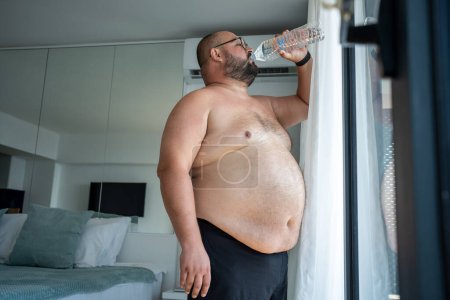 Téléchargez les photos : Le gros souffre de mourir de chaleur. barbu obèse gars boit beaucoup d'eau debout à la fenêtre ouverte à côté de climatiseur essayant de faire face à la chaleur anormale de l'été sueurs reconstitue fortement l'équilibre de l'eau - en image libre de droit