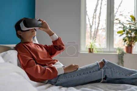 Téléchargez les photos : Adolescente détendue enfant dans des lunettes vr regarder 360 degrés vidéo éducative immersive. Femme en lunettes VR utilisant casque métaverse couché sur le lit à la maison. Réalité virtuelle, enfants et éducation - en image libre de droit