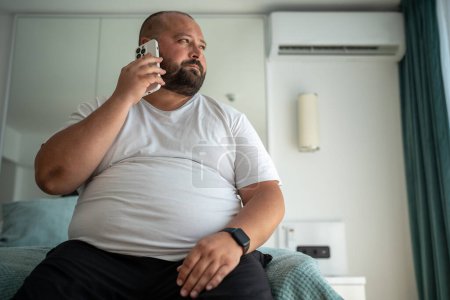 Photo for Fat guy calls on phone, sitting on bed and looking out window. Extra over obese bald bearded guy makes business call, signs up for gym to put figure appearance in order, to love accept body. - Royalty Free Image