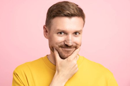 Photo for Portrait of playful man with sly insidious look touching chin by hand looking at camera isolated on pink studio background. Bearded smiling guy flirting, dodgy shifty slippery person for advertising. - Royalty Free Image