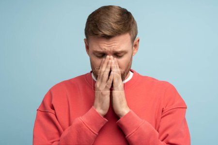 Photo for Portrait of frustrated feeling desperate man closing face with hands and eyes on blue background. Guy living through loss of loved one, heartbreaking, divorce or death of life partner. Emotional pain. - Royalty Free Image