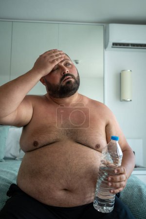 Téléchargez les photos : Gros homme avec bouteille d'eau à la main souffrant de chaleur dans la chambre d'hôtel en vacances par temps chaud d'été. Surpoids barbu chauve d'âge moyen mâle a des maux de tête, hypertension artérielle, problèmes de santé. - en image libre de droit