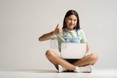 Photo for Teen girl with laptop in hands showing thumb up sitting on gray background looking at camera. Online, distant remote education, educational university high school college program, advertising banner. - Royalty Free Image