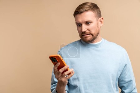 Photo for Scared frustrated man reding message in smartphone looking at screen isolated on beige background. Bearded middle aged guy having bad news, problems, troubles. Male feeling confusion, desperation. - Royalty Free Image