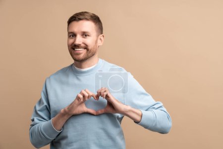 Photo for Smiling man showing heart hand gesture on chest on beige background looking at camera. Love romantic sign symbol, share support, kindness concept. Banner, poster for advertisement with copy space. - Royalty Free Image