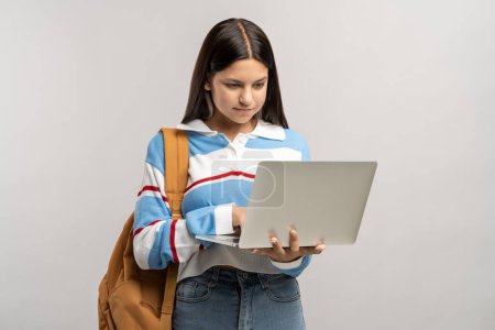 Photo for Interested student teen girl with backpack looking at laptop screen on gray background. Focused teenager busy learning studies, studying online. Education in college, high school, university concept. - Royalty Free Image