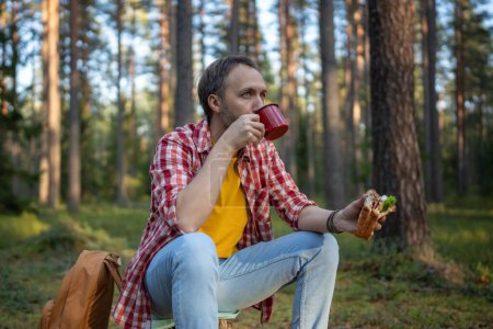 Téléchargez les photos : Homme randonneur boire du thé de tasse pendant la randonnée dans la forêt. Homme pendant une petite halte manger un sandwich et boire du café dans les bois d'automne, se reposer arrêté pour une pause après une promenade dans les bois. - en image libre de droit