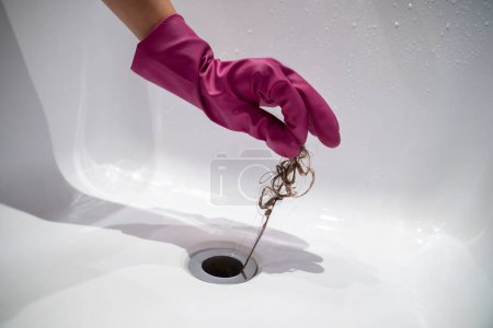 Hand of woman in pink rubber glove taking of hair clump from water drain, blocked pipe in bath. Woman cleaning sewer trap. Bath plug hole blocked with dirt. Problem with shower sewerage in bathroom.