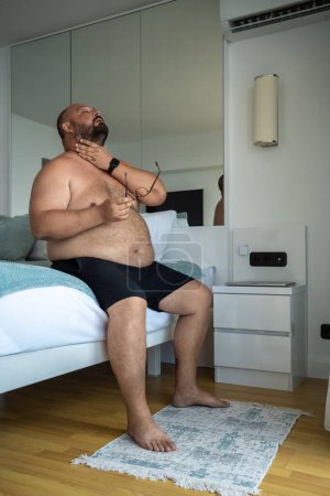 Photo for Overweight bearded man in shorts in an air-conditioned room escaping from heat in hotel on vacation rubbing neck holding eyeglasses. Obesity male, health problems, plump chubby guy suffer from hot. - Royalty Free Image