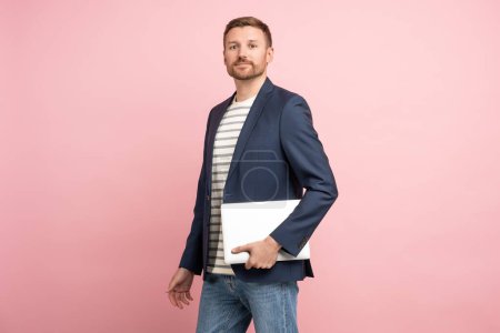 Photo for Man trader, crypto investor, entrepreneur, financier with laptop in hands on pink background. Bearded well groomed smiling guy wearing jacket, jeans looking at camera on advertisement banner poster. - Royalty Free Image