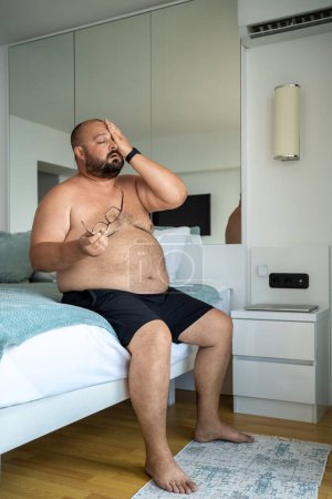 Photo for Fat man wipes sweat from face sitting on bed of hotel room. Chubby tormented suffering man hardly tolerate exorbitant summer heat, sits under air conditioner, uses all available means for cooling. - Royalty Free Image