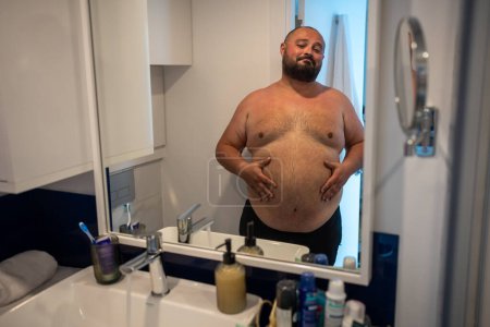 Foto de Hombre gordo mirando el espejo sonriendo tocando gran abdomen en el baño en casa. Tipo barbudo insatisfecho con la forma de su cuerpo. Obesidad, sobrepeso masculino, peso extra, problemas saludables, control de peso. - Imagen libre de derechos