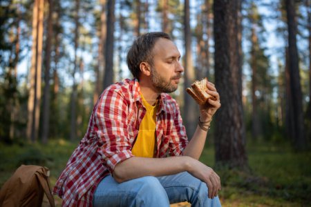 Photo for Carefree man eating sandwich snack during hike walk travel camping vacation, feels calm relax in nature landscape. Happy hiking male resting taking break with healthy food in forest. Autumn nature. - Royalty Free Image
