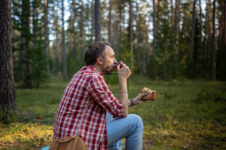 Foto de Hombre excursionista beber té de taza durante el senderismo en el bosque. Masculino durante un pequeño descanso comiendo sándwich y tomando café en el bosque de otoño, descansando detenido para descansar después de caminar en el bosque. - Imagen libre de derechos