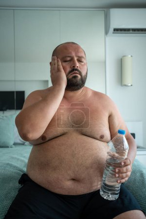 Téléchargez les photos : Gros homme avec bouteille d'eau à la main souffrant de chaleur dans la chambre d'hôtel en vacances par temps chaud d'été. Surpoids barbu chauve d'âge moyen mâle a des maux de tête, hypertension artérielle, problèmes de santé. - en image libre de droit