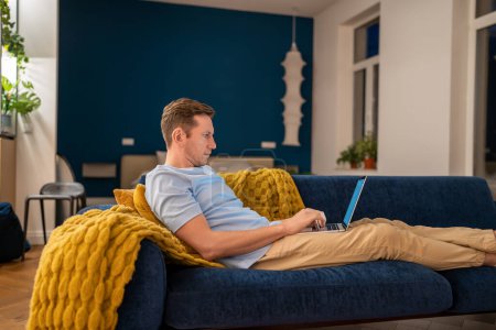 Photo for Man sit on couch with laptop on knees. Focused serious unsmiling young guy working remotely online from home lying on sofa with computer, studying, relaxing, chilling, browsing Internet, checking news - Royalty Free Image