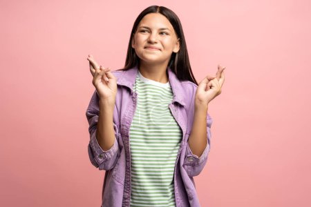 Photo for Teenage girl smiling dreaming with fingers crossed. Brunette schoolgirl hopes for good luck, folding hands in happy gesture. Teenager worried, anticipating victory. Isolated on pink studio background. - Royalty Free Image