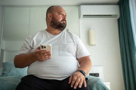 Photo for Obese unshaved man with phone and fitness tracker looking at window. Overweight middle-aged man thinks on weight reduction, diet and calories calculation. Weight control and healthy food concept - Royalty Free Image