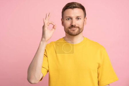 Photo for Man raises hand in gesture ok. Light smile middle aged guy in yellow T-shirt agreeing assenting shows generally arm sign, connecting index finger thumb of palm. Isolated portrait studio background. - Royalty Free Image
