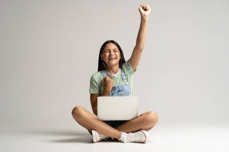 Photo for Happy smiling teen girl rejoycing sitting on gray background with laptop on legs raising hand up showing yes gesture. Overjoyed emotional teenager student having good excellent marks studying online. - Royalty Free Image