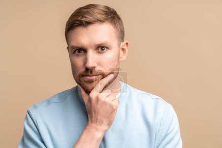 Photo for Pensive puzzled thoughtful man looking at camera touching chin isolated on studio beige background. Embarrassed guy feeling confusion solves problems, struggling with choice. Male pondering idea. - Royalty Free Image