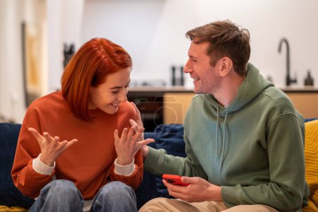 Photo for Happy overjoyed couple man woman reading good news in smartphone sitting on couch at home. Wife husband smiling rejoicing together, gesturing hands, having amazed wondered faces. - Royalty Free Image