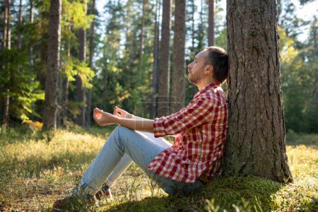 Foto de Hombre sentado bajo un pino en el bosque con los ojos cerrados meditando en unidad con la naturaleza en un día soleado. Chico relajante descansando sintiendo paz, tranquilidad. Meditación, concepto de experiencia de práctica espiritual. - Imagen libre de derechos